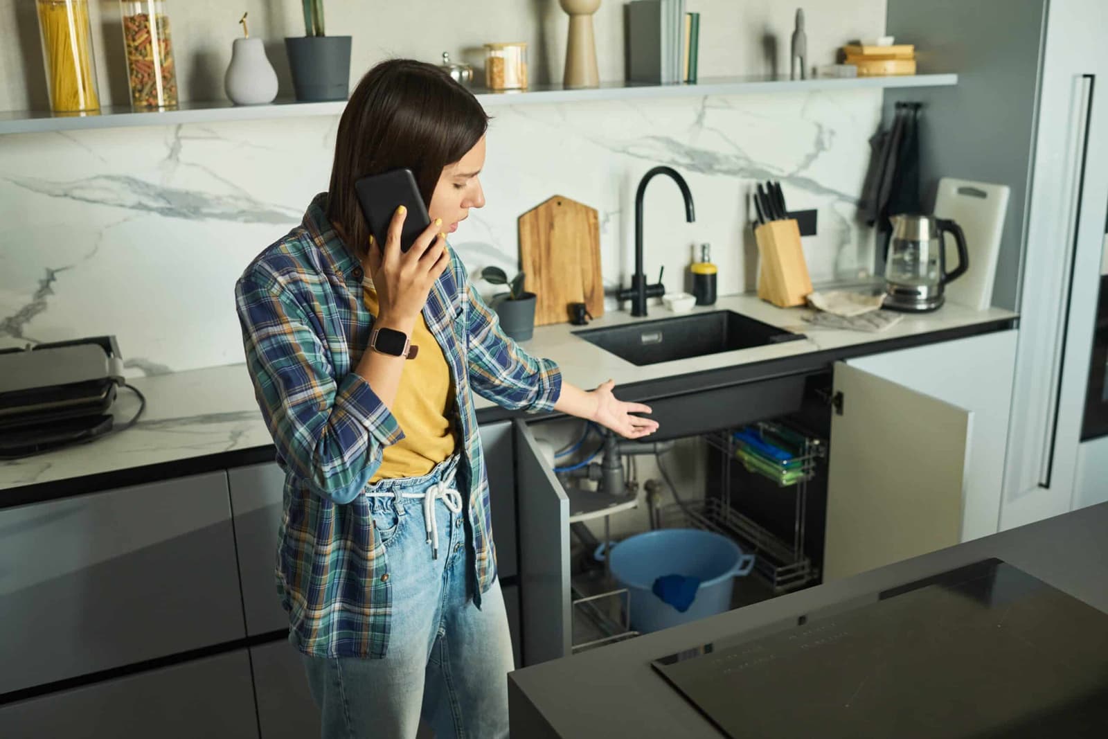 Woman on the phone crouching beside an open cupboard under a kitchen sink