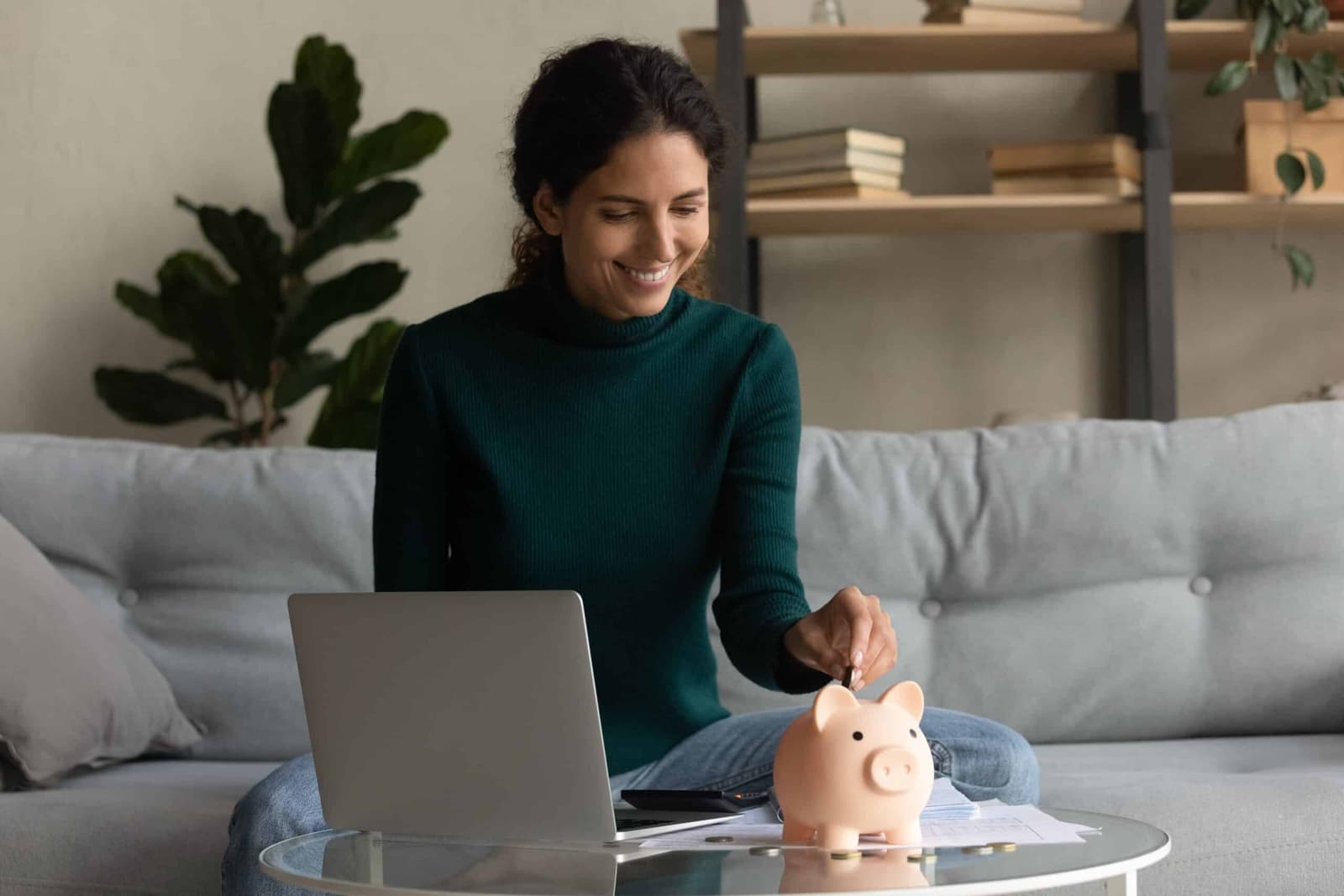 Woman on a sofa with a laptop putting a coin into a piggy bank
